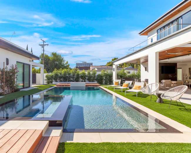 The image shows a modern pool area with a rectangular swimming pool surrounded by wooden decking and lounge chairs, adjacent to a stylish house with open outdoor living spaces.