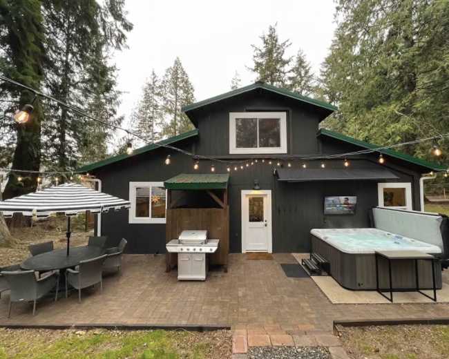 The image shows a modern cabin with a barbecue grill and a hot tub on a patio, surrounded by trees.