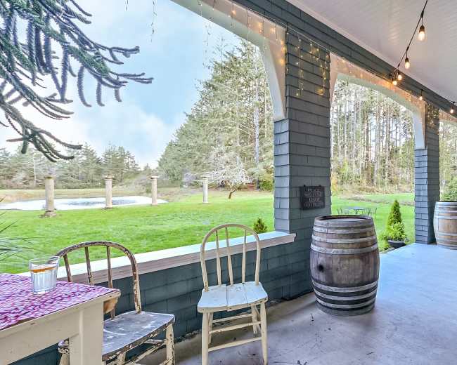 A porch with two wooden chairs and wine barrels, decorated with string lights, overlooking a green lawn and trees in the background.