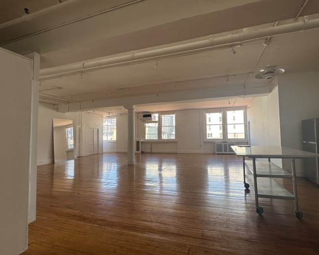 The image shows a spacious, empty room with wooden flooring, large windows, and minimalist furnishings.