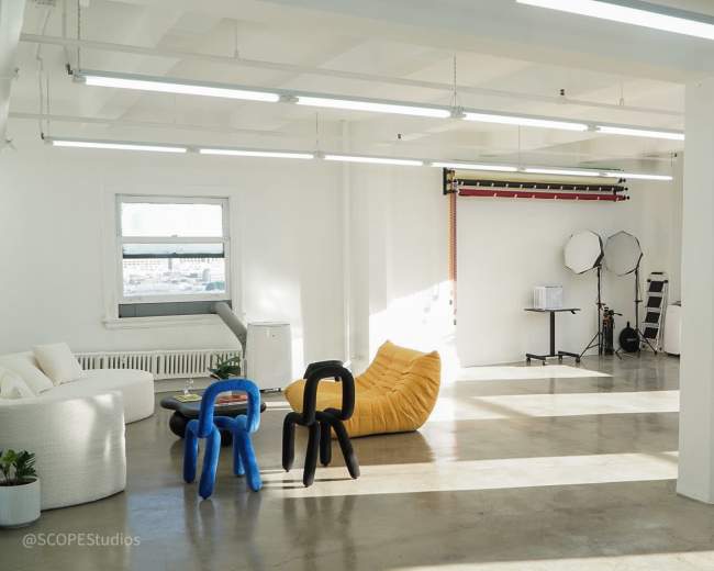 The image shows a spacious, well-lit studio with modern furniture, including a white couch, a yellow bean bag, and two colorful chairs, alongside photography equipment and large windows.