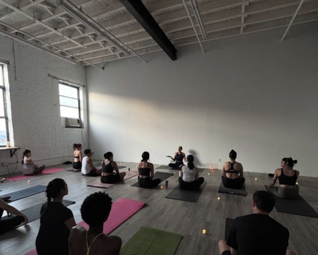 A group of people sits on yoga mats in a dimly lit room, facing an instructor seated at the front, with candles placed around them.