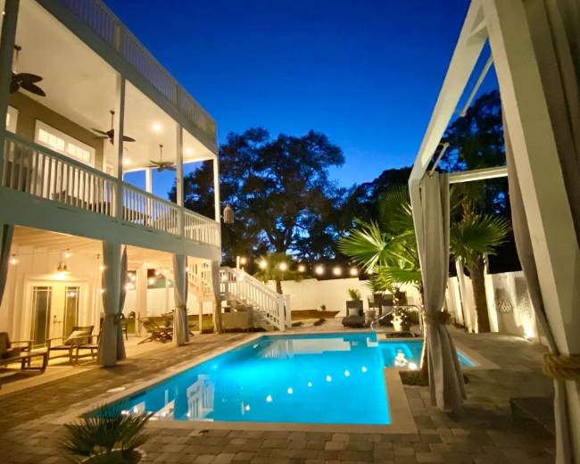The image shows a modern house with a swimming pool lit up at night, surrounded by palm trees and outdoor seating areas.