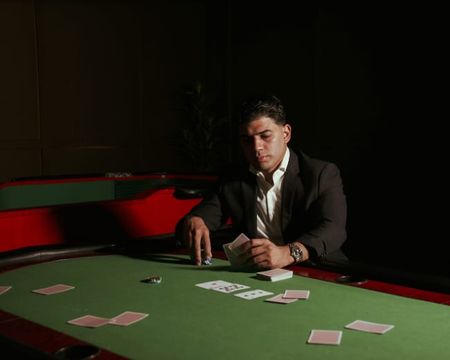 A man in a black suit sits at a green felt poker table, examining a set of playing cards with a focused expression.