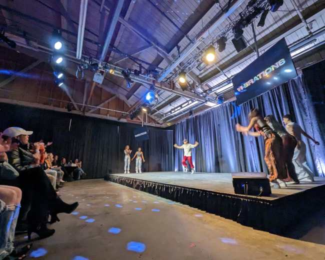 A group of dancers performs on a stage while an audience watches from the side.