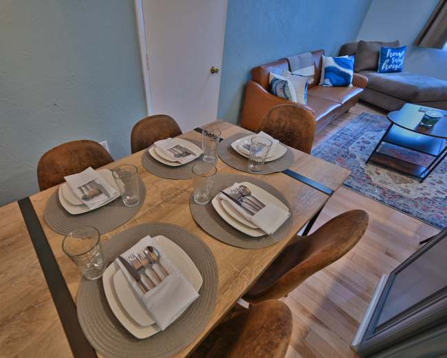 A dining table is set with plates, glasses, and utensils, next to a living area featuring a sofa and a rug.