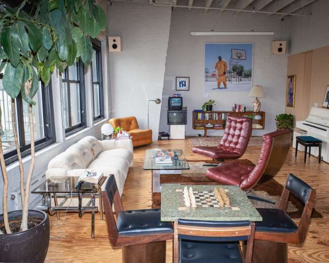 The image shows a spacious living room featuring a mix of seating including a beige sofa, red lounge chairs, and a wooden table with a chessboard, surrounded by houseplants and artwork on the walls.