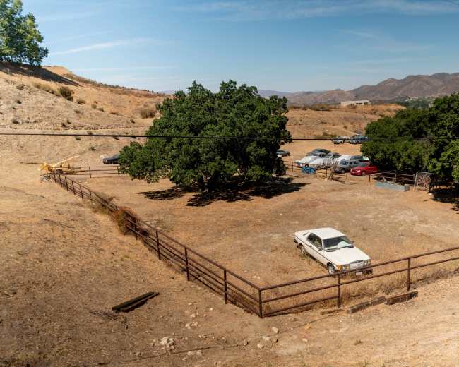 A fenced dirt lot contains an old white car surrounded by sparse vegetation and distant mountains.
