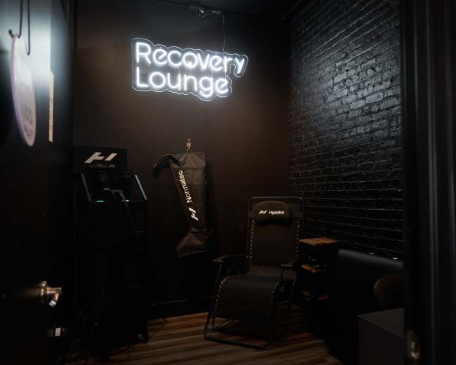 The image shows a dimly lit room with a neon sign reading "Recovery Lounge," a black brick wall, a reclining chair, and a recovery machine.