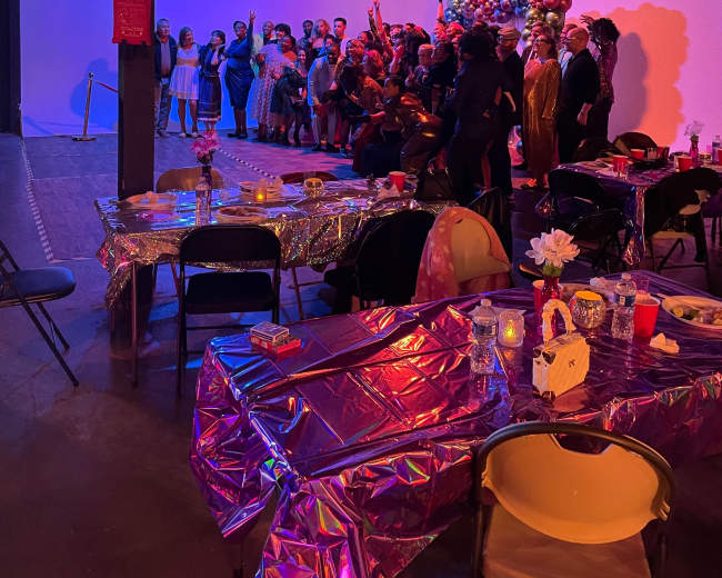 A large group of people gathers for a celebration in a dimly lit room decorated with balloons and festive tables.
