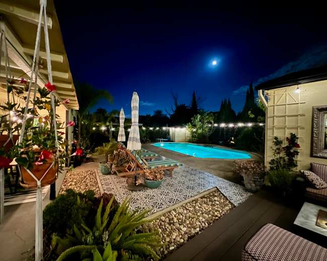 The scene features a well-lit backyard at night with a swimming pool, decorative landscaping, and a clear moon in the sky.
