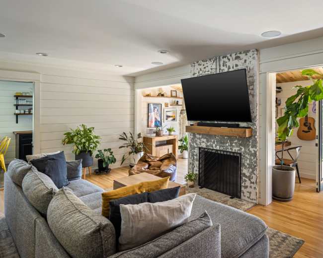 A modern living room features a large sofa, a mounted television above a fireplace, and an open layout leading to a dining area and a porch with plants and a guitar.