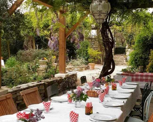 A long outdoor dining table is set with plates and checkered napkins beneath a pergola, surrounded by lush greenery and blooming wisteria.