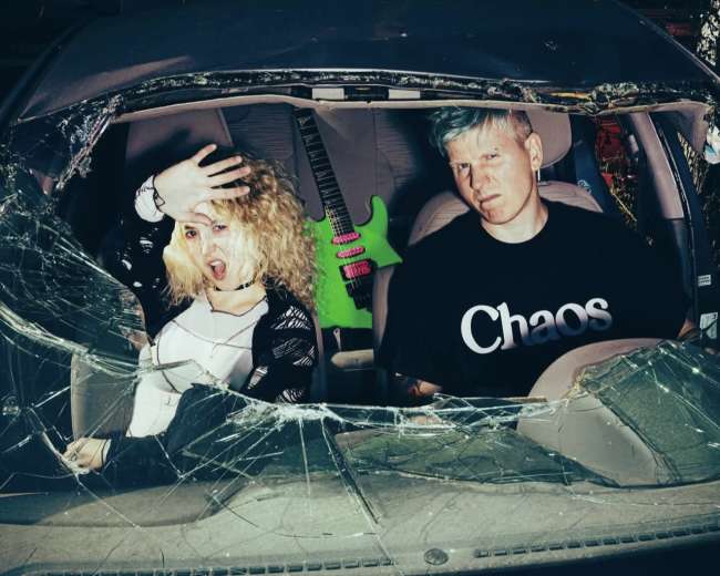 A woman with curly blonde hair and a man with blue hair sit inside a car with a shattered windshield, and a green guitar is visible in the background.