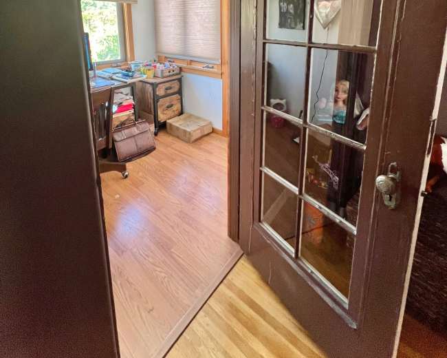 The image shows an open door leading into a brightly lit room with wooden floors, a desk cluttered with items, and a window with sheer shades.