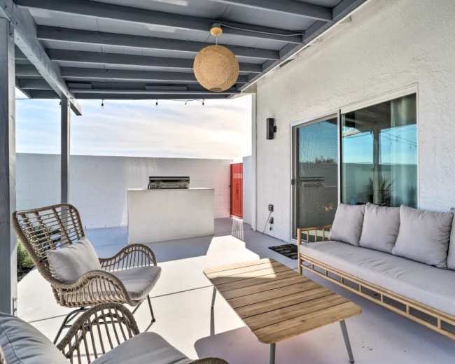 The image depicts a covered patio area with a rattan sofa, two chairs, a wooden coffee table, and a grill in the background.