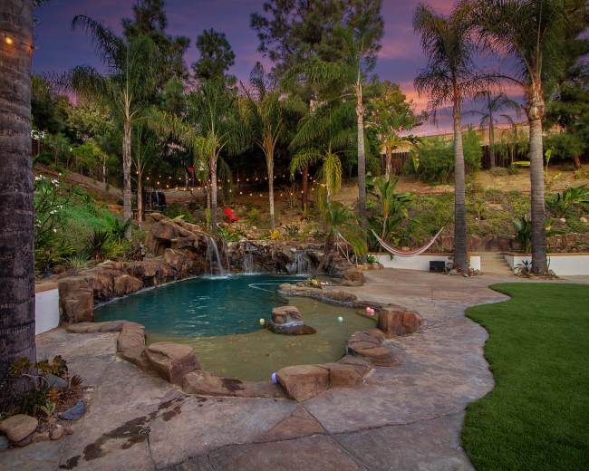 A landscaped backyard featuring a swimming pool with a rock waterfall, surrounded by palm trees and decorative lighting.