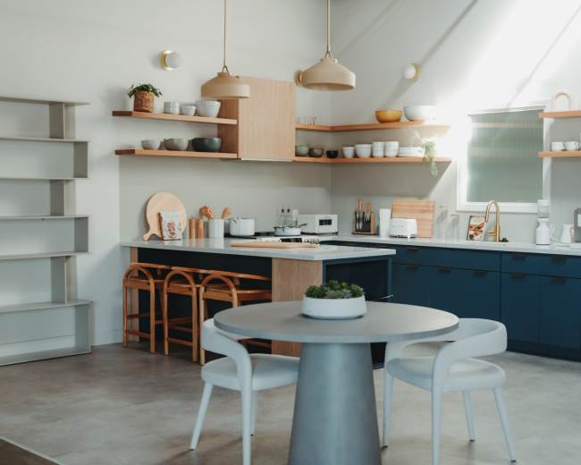 A modern kitchen features blue cabinetry, open shelving with dishware, and a circular dining table with white chairs.