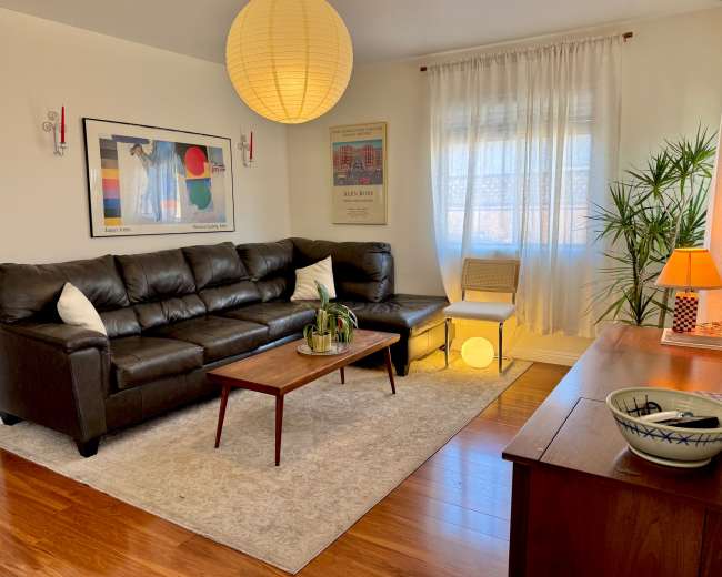The image shows a well-lit living room featuring a black leather sectional sofa, a wooden coffee table, a rug, and decorative plants.