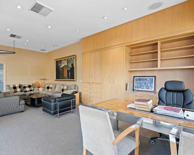 The image shows an office space featuring a desk, a black leather chair, a seating area with a couch, and wooden cabinetry.