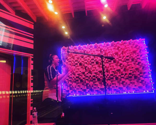 A person is recording vocals in a sound studio with colorful lighting and acoustic wall paneling.