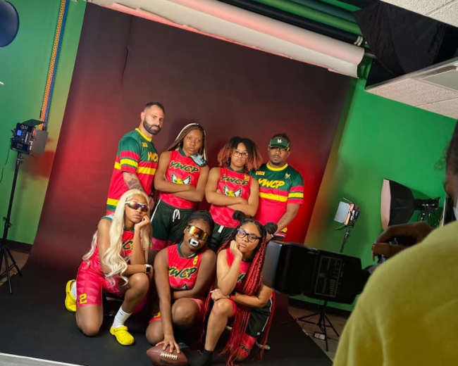 A diverse group of people poses in a studio setting, wearing colorful outfits and sports gear against a black backdrop with red lighting.