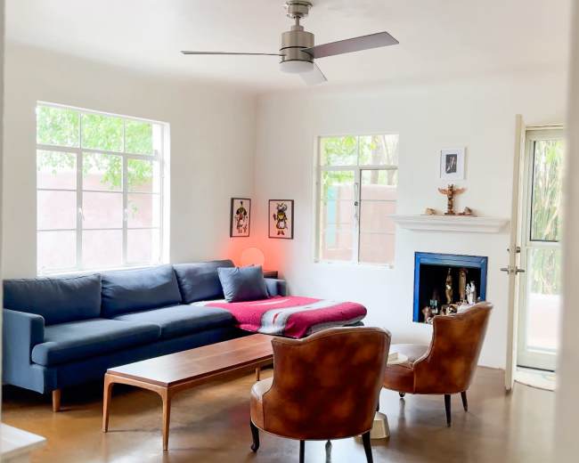 The image shows a living room featuring a blue sofa, a wooden coffee table, two brown chairs, and large windows that allow natural light to fill the space.