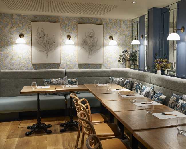 The image shows a modern restaurant interior featuring a long wooden table with stationery, surrounded by upholstered seating and decorative floral wallpaper.