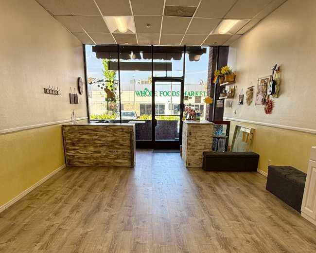 The image depicts a spacious, well-lit retail space with wooden flooring, a reception area on the left, and large windows facing a street with a view of a Whole Foods Market.