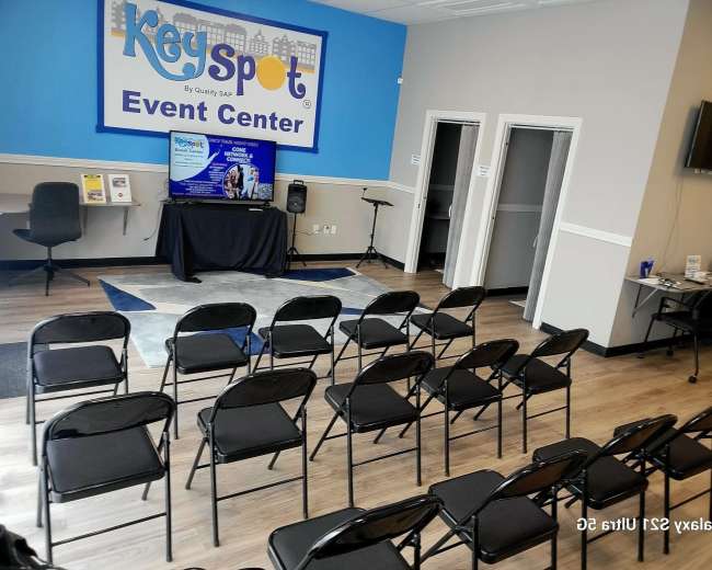 The image shows a semi-empty event center with rows of black folding chairs facing a television screen displaying event information.