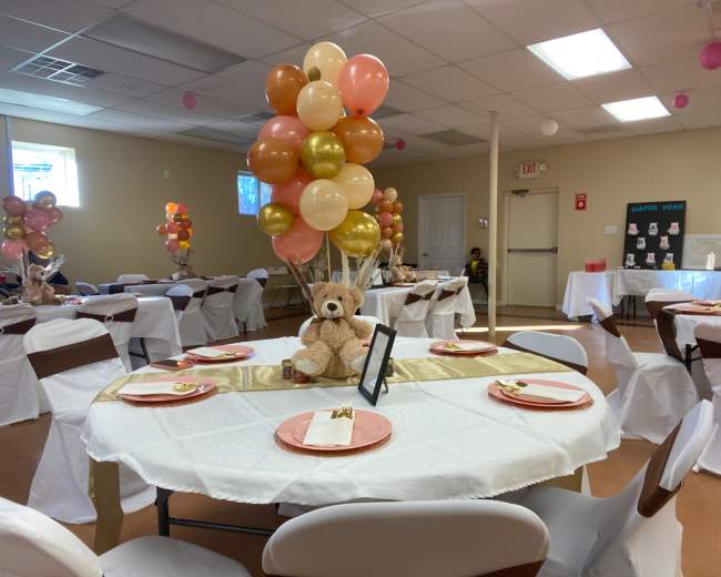 The scene shows a decorated party venue with tables set with white tablecloths, colorful balloon centerpieces, and a teddy bear at the center of each table.