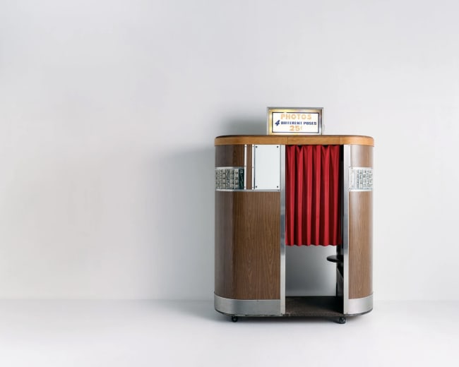 A vintage photo booth with a wooden exterior and red curtains stands against a plain wall.