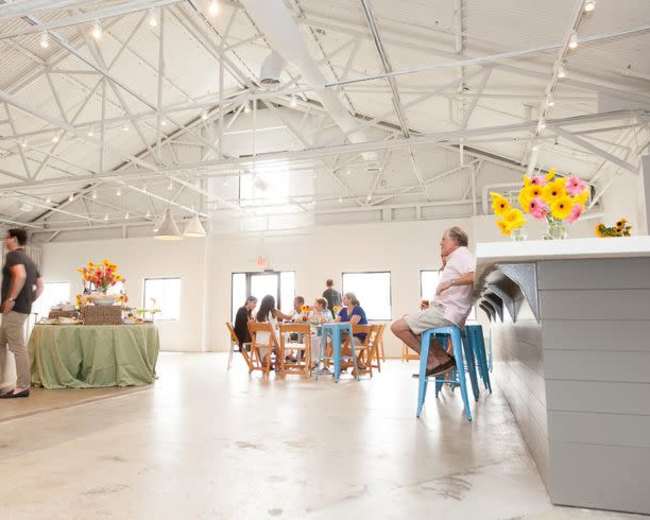 A spacious event hall features a long bar, a dining area with wooden tables, and colorful flower arrangements.