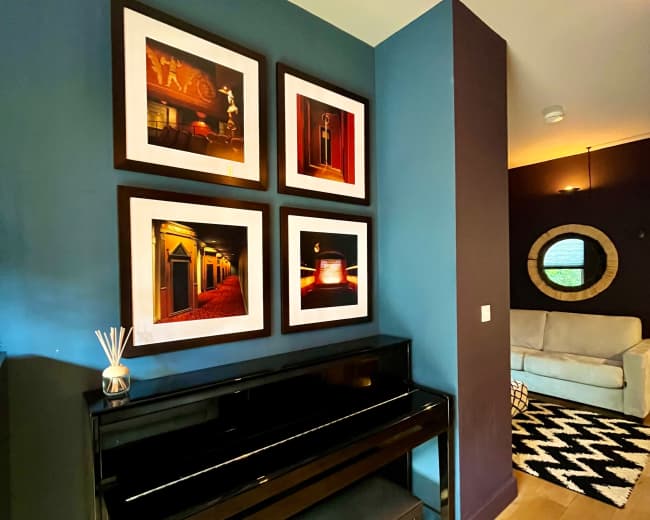 A black piano is positioned against a dark blue wall, adorned with four framed photographs, with a cozy seating area visible in the background.