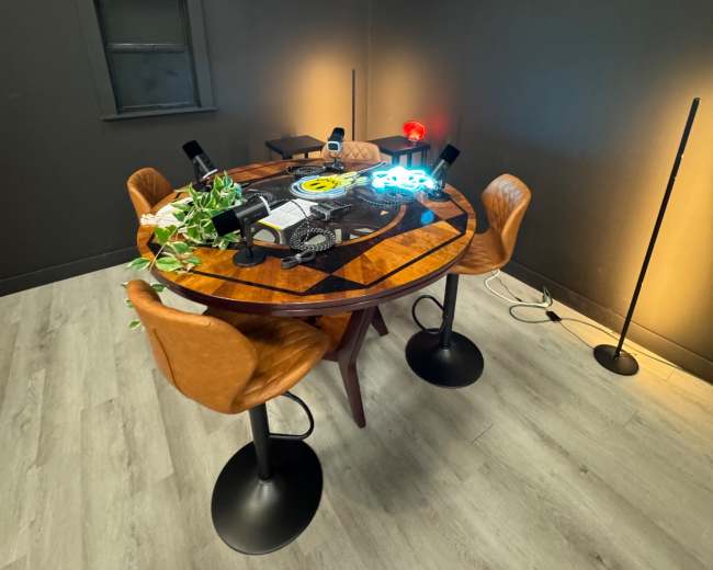 A round wooden table with a game board is set in a dimly lit room, surrounded by two chairs and a floor lamp.