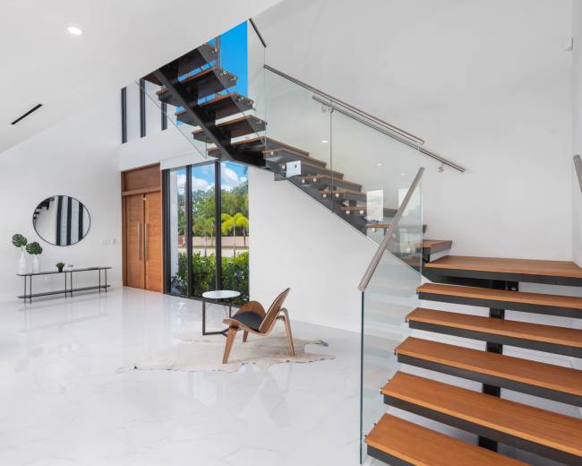 The image shows a modern interior featuring a staircase with wooden steps and glass railings, alongside a sleek coffee table and a round mirror on the wall.