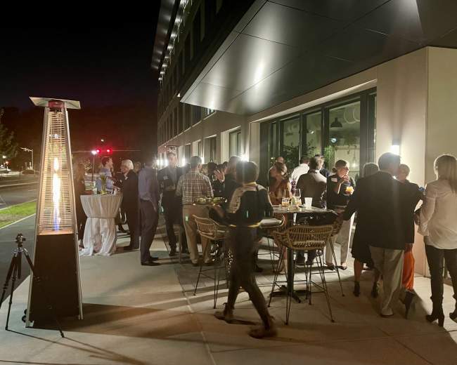 A group of people socialize at an outdoor gathering next to a modern building, illuminated by gas heaters and ambient lighting.