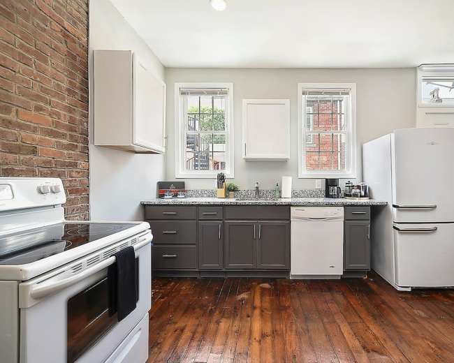 The kitchen features a combination of brick and painted walls, with a stove, refrigerator, and dishwasher arranged along a countertop.