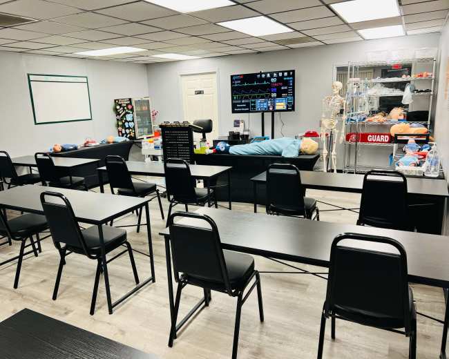 A classroom is arranged with black tables and chairs, featuring medical training supplies and a monitor displaying health data.