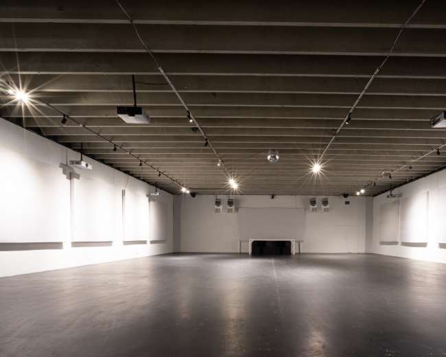 The image shows a spacious, empty room with high ceilings, polished black flooring, and projector equipment mounted on the walls.