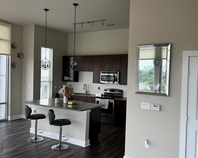 The image shows a modern kitchen and dining area featuring dark wood cabinets, stainless steel appliances, and two barstools at a counter.