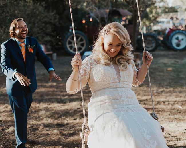 A woman in a wedding dress swings joyfully while a man in a suit playfully approaches her in a rustic outdoor setting.