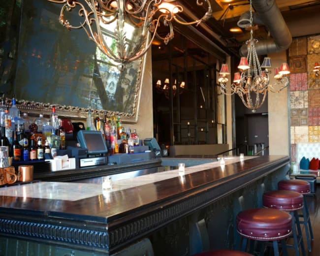 The image shows a stylish bar with a long counter, ornate chandeliers, and a decorative wall featuring colored tiles.