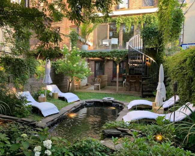The image shows a backyard oasis with a small pond surrounded by lounge chairs, lush greenery, and a spiral staircase leading to a balcony.