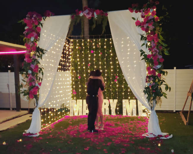 A couple embraces in front of a floral arch adorned with lights and a "MARRY ME" sign, surrounded by rose petals and candles on a grassy area.