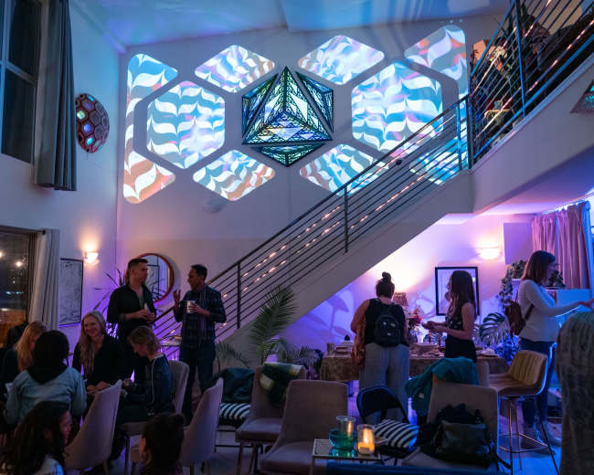 The interior of a modern venue features a staircase adorned with geometric light projections, while guests engage in conversation across various seating areas.