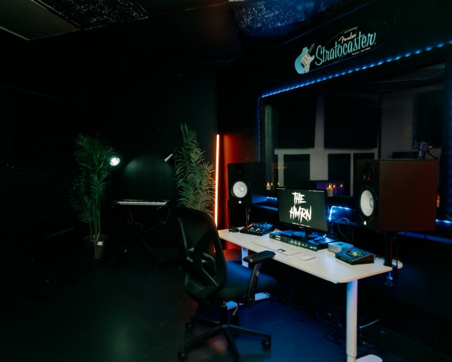 The image shows a music production studio featuring a desk with audio equipment, speakers, and a computer monitor, alongside a keyboard and decorative plants.