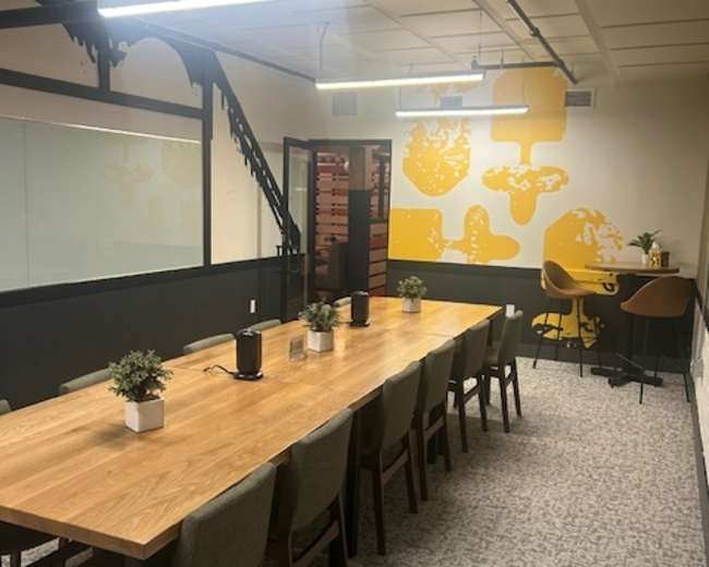 The image shows a modern conference room with a long wooden table surrounded by green chairs, and a yellow accent wall featuring abstract designs.