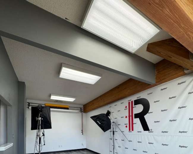 The image shows a well-lit photo studio with two lighting setups, a backdrop featuring the letter "R," and a spacious wooden-beamed ceiling.
