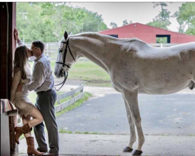 A couple kisses near the entrance of a stable while a white horse stands nearby.